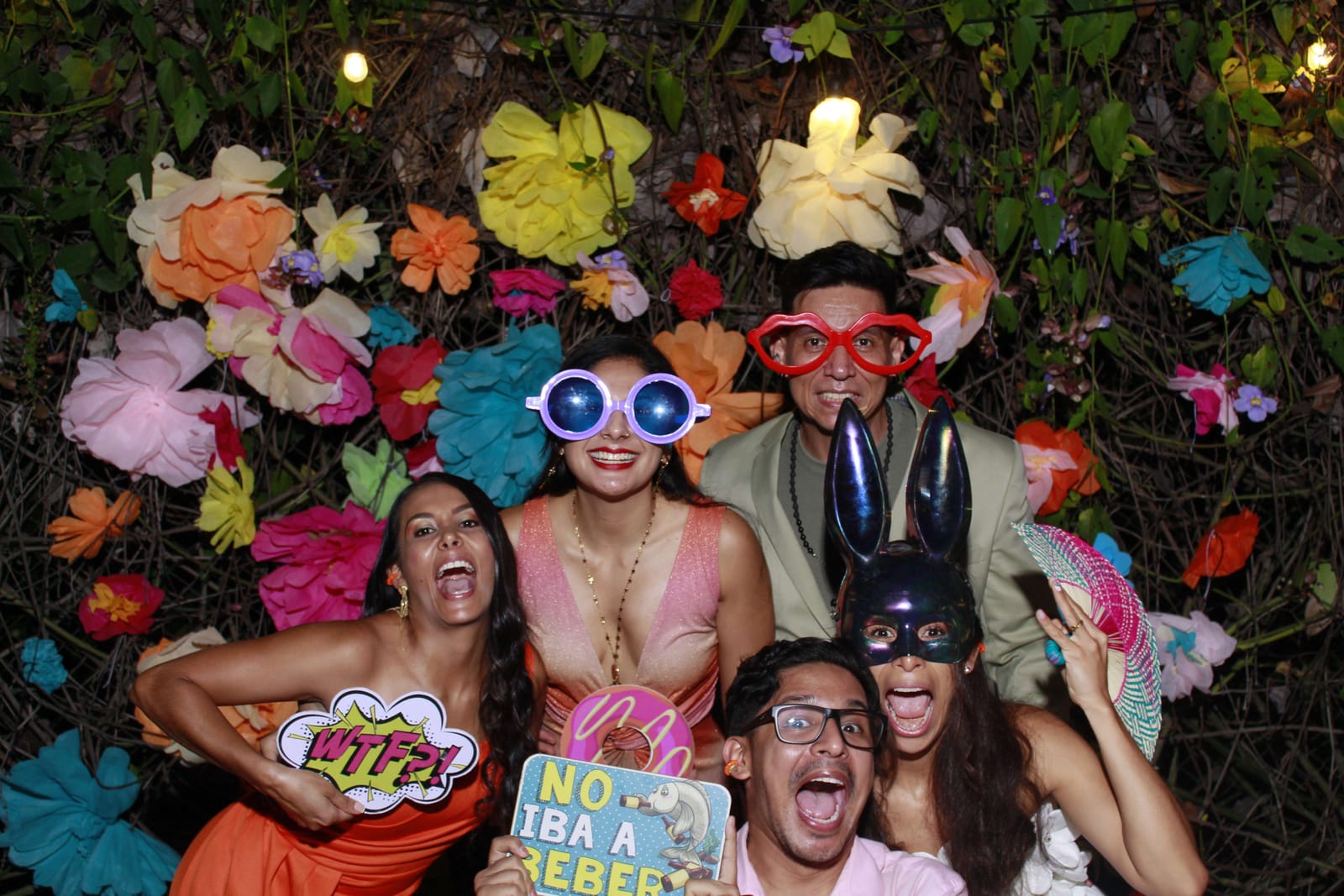 Cinco invitados posando con gafas y antifaces frente al fondo floral de una cabina de fotos de Kuva